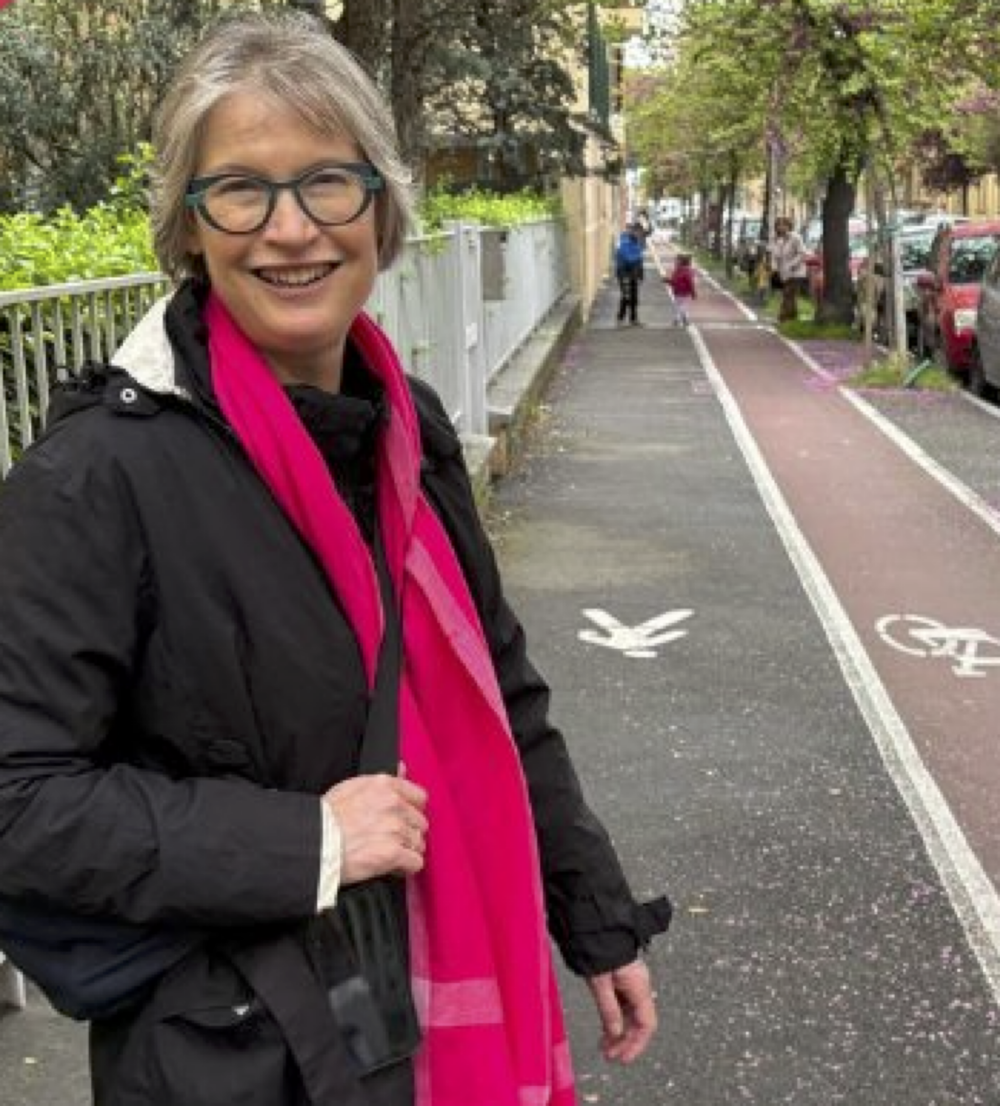 An image of Dr Anna Stevenson wearing a black coat and pink scarf, with a grey bob and glasses walking on a split pedestrian and cycling path.