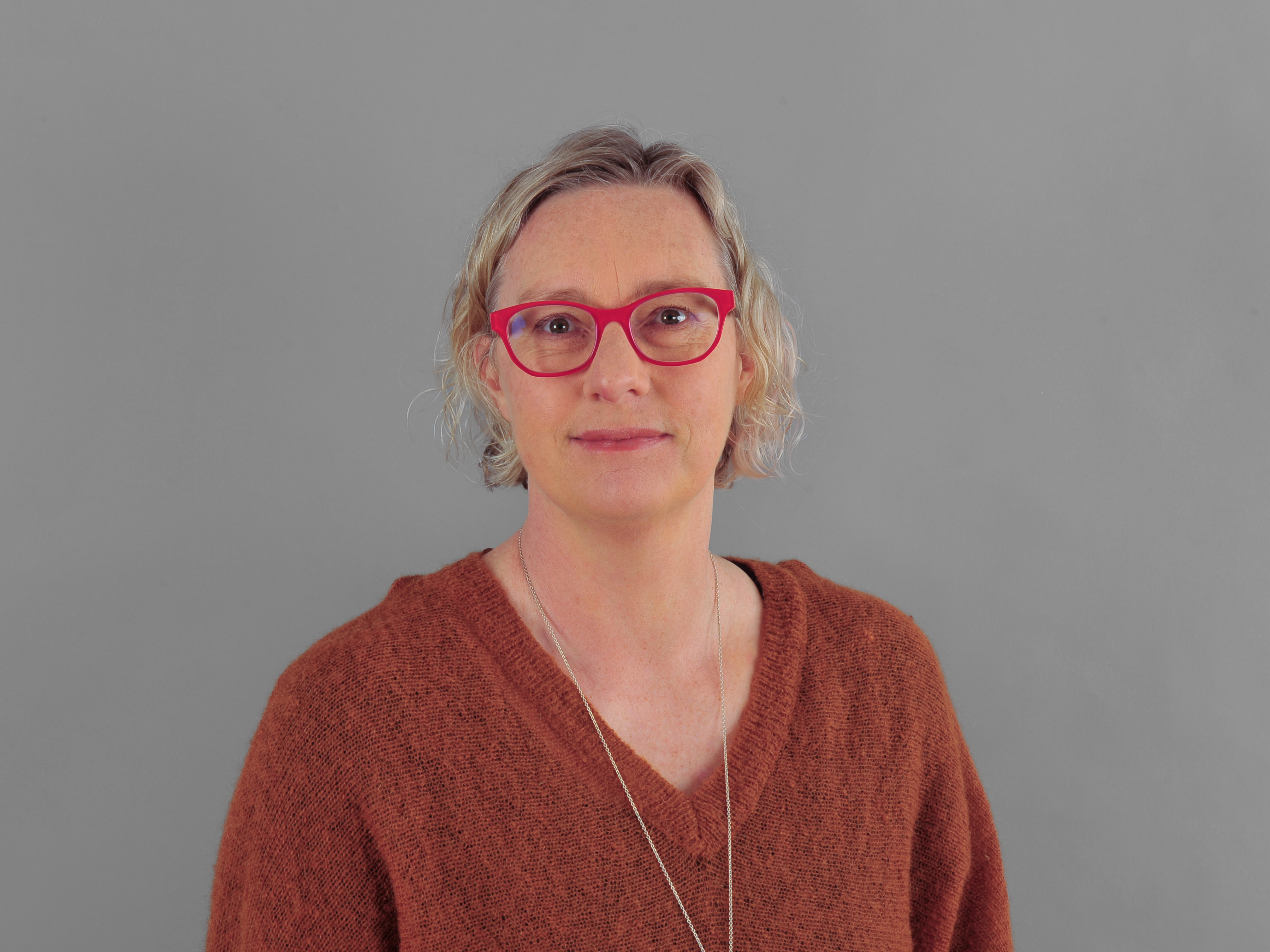 A headshot of Dr Caroline Shaw with a blond bob, brown eyes, wearing red glasses and an orange longsleeve jumper.
