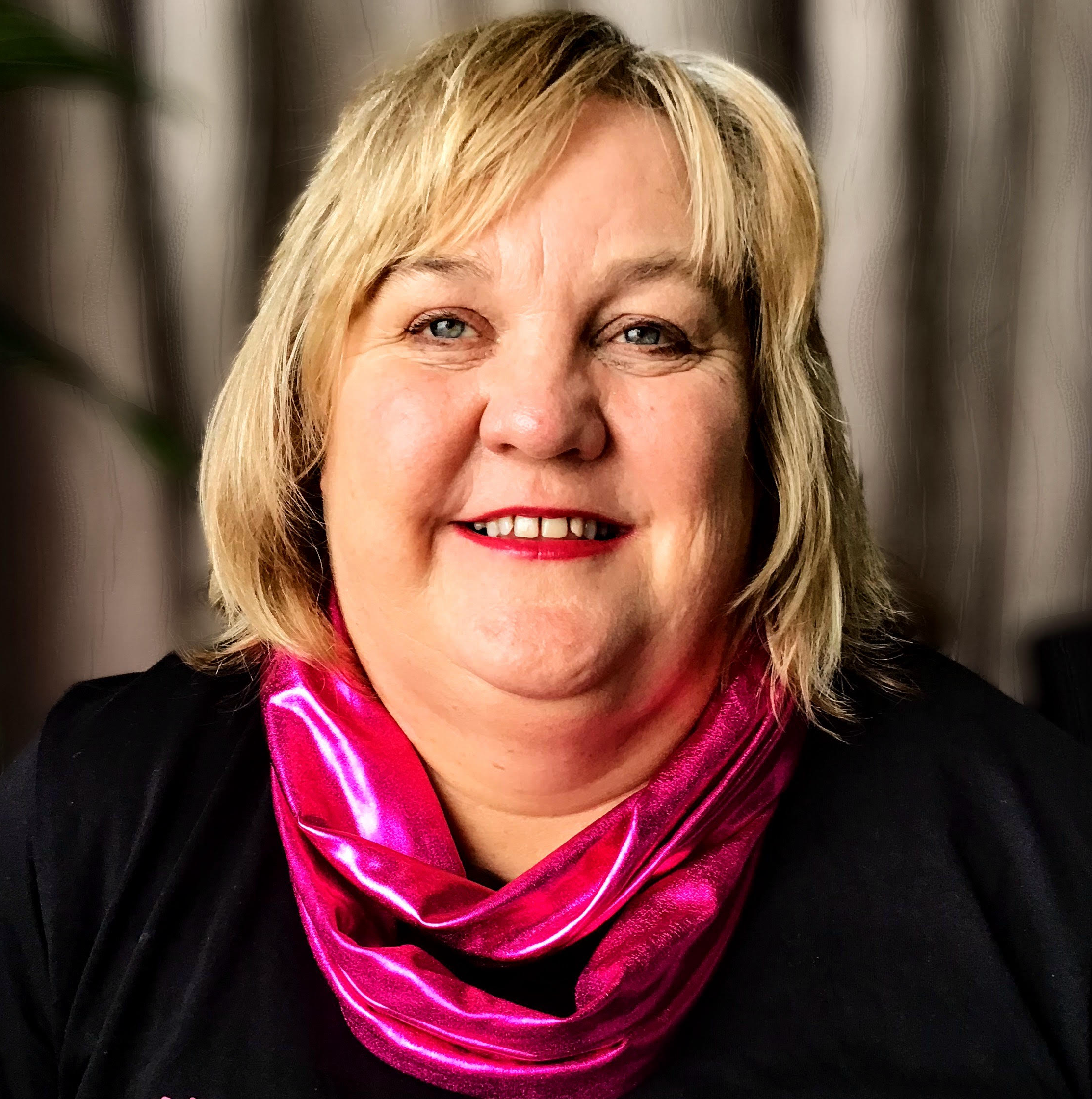 An image of Julie Woods with blond hair, blue eyes, wearing a black shirt and pink scarf.