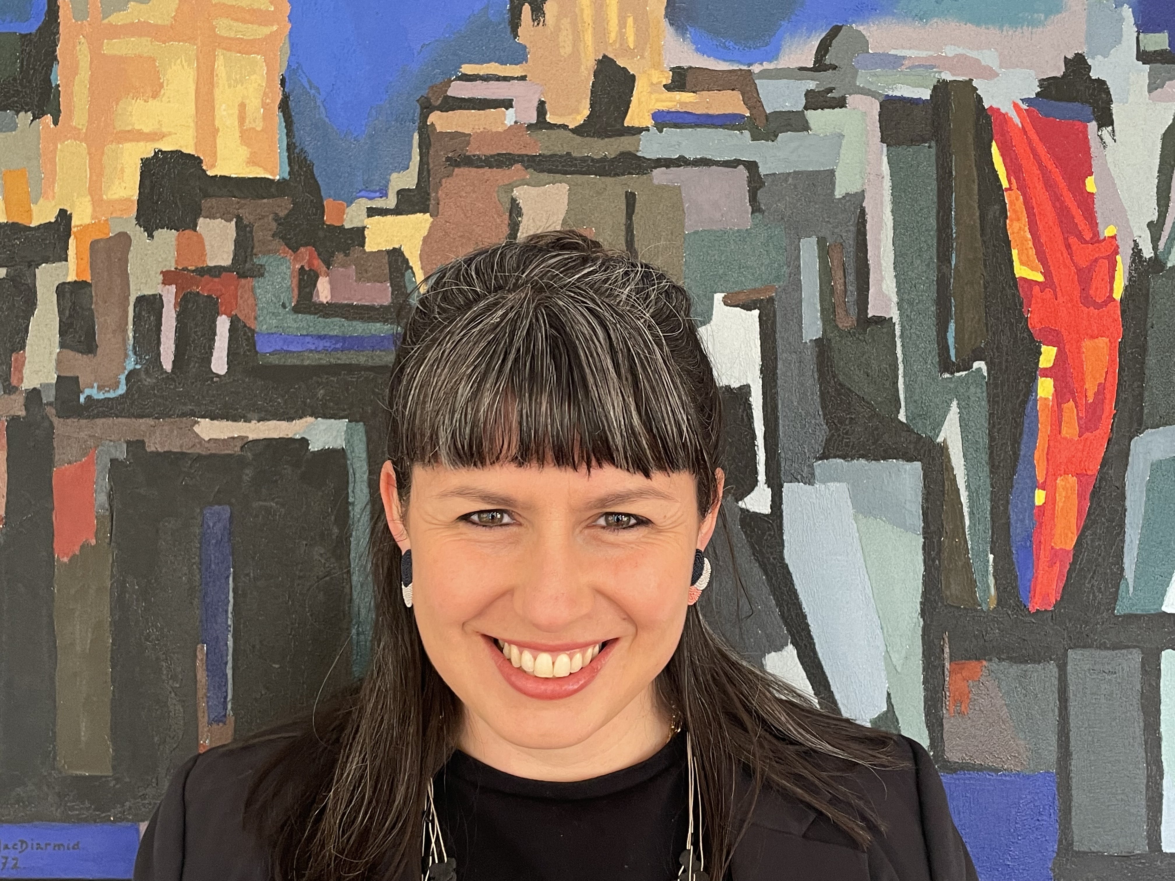 A headshot of Vida Christeller with salt and pepper hair, blue eyes, wearing a black longsleeve with a necklace in front of a mural.